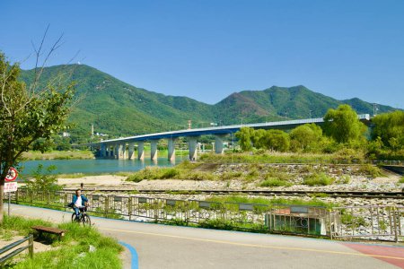 Photo for Hanam City, South Korea - October 1, 2023: A cyclist pedals along a bike path, with Deokpung Stream in the foreground and Paldang Bridge and Yebongsan Mountain in the distance. - Royalty Free Image