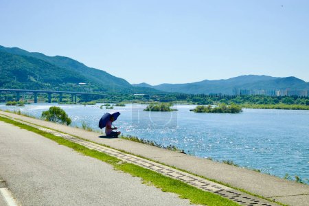 Photo for Namyangju City, South Korea - September 30, 2023: A solitary figure with an umbrella sits by the Han River, contemplating the serene view of small islets, Paldang Bridge, and Hanam City. - Royalty Free Image