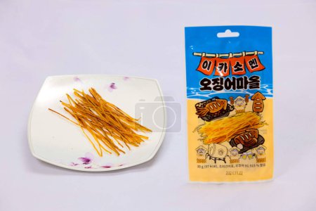 Photo for Ulsan, South Korea - January 14, 2024: A display of dried squid in long thin strips on a plate, alongside its original "Squid Village" packaging, showcasing a traditional Korean seafood snack against a white background. - Royalty Free Image