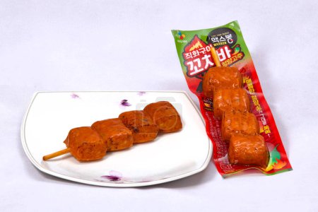 Photo for Ulsan, South Korea - January 14, 2024: A fiery spicy hot bar, with one still in its packaging and another served on a plate, featuring four chunks of meat on a stick, set against a white background, highlighting this intensely spicy Korean snack. - Royalty Free Image