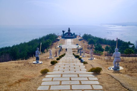 Foto de Samcheok City, Corea del Sur - 28 de diciembre de 2023: Vista desde lo alto de una colina, con un camino que conduce a la escultura Lady Suro, flanqueada por estatuas de soldados y el Mar del Este en el fondo. - Imagen libre de derechos