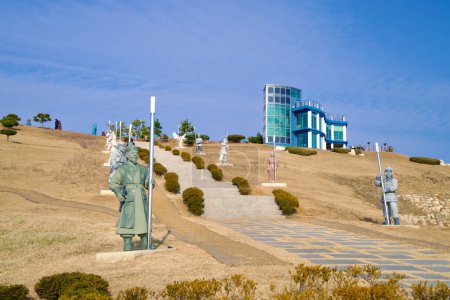 Foto de Samcheok City, Corea del Sur - 28 de diciembre de 2023: Escaleras que conducen a un edificio del observatorio en lo alto de una colina en Lady Suro Memorial Park, alineado con estatuas de soldados de la era Silla junto a céspedes ondulados. - Imagen libre de derechos