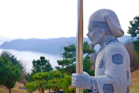 Téléchargez les photos : Samcheok City, Corée du Sud - 28 décembre 2023 : Sculpture en pierre d'un érudit de l'ère Silla tenant un poteau métallique dans le parc commémoratif Lady Suro, avec des arbres, la mer de l'Est et le port d'Imwon en arrière-plan. - en image libre de droit