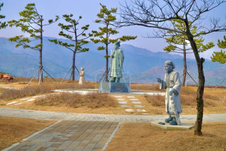 Foto de Samcheok City, Corea del Sur - 28 de diciembre de 2023: Esculturas de hombres y eruditos de la era de Silla de pie en una colina en el Parque Memorial Lady Suro, enmarcado por árboles y montañas en el fondo. - Imagen libre de derechos