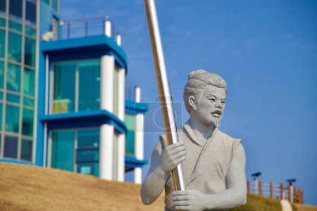 Foto de Samcheok City, Corea del Sur - 28 de diciembre de 2023: Una escultura de soldado de la era Silla colocada contra un cielo azul, con el edificio del observatorio del Lady Suro Memorial Park al fondo. - Imagen libre de derechos