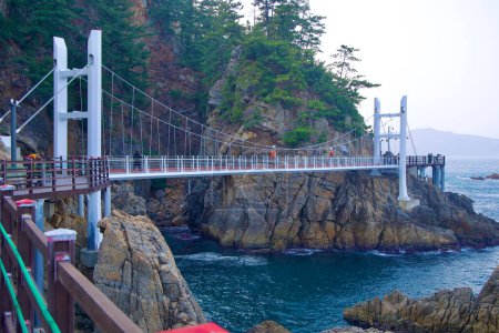 Foto de Samcheok City, Corea del Sur - 28 de diciembre de 2023: Un tiro ancho captura el puente colgante en el Sendero de Senderismo de la Cueva de Chogok Yonggul, mostrando su envergadura completa a través de la costa rocosa contra el telón de fondo del Mar del Este. - Imagen libre de derechos