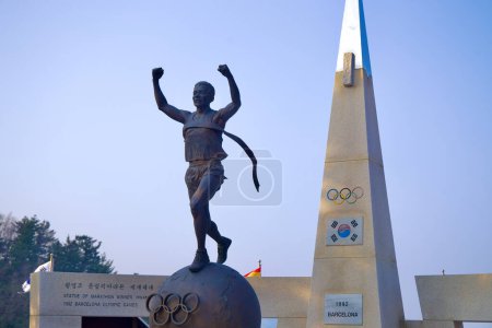 Foto de Samcheok City, Corea del Sur - 28 de diciembre de 2023: Primer plano de la estatua de Hwang Young-jo, que lo representa corriendo a través de la cinta de la línea de meta en una pelota con anillos olímpicos, una pirámide alta, bandera coreana y anillos olímpicos en el fondo. - Imagen libre de derechos