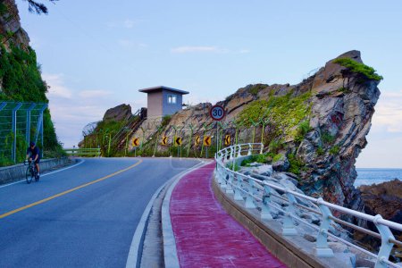 Photo for Gangneung City, South Korea - July 29th, 2019: A tall rocky outcrop along Heonhwa Road features a military lookout post, with a cyclist navigating the curvy coastal road under a blue sky, embodying the route's blend of natural beauty and strategic im - Royalty Free Image