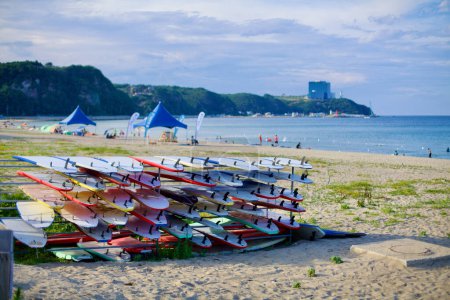 Photo for Gangneung City, South Korea - July 29th, 2019: Surfboards stacked on racks on the sand at Geumjin Beach, with the East Sea waters and Tops 10 Hotel & Resort visible on the seaside hills. Overhead tarps provide shade for beachgoers, with people enjoyi - Royalty Free Image