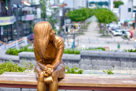 Gangneung City, South Korea - July 29th, 2019: A golden statue of a young woman sits on a park bench overlooking Wolhwa Street in downtown Gangneung, depicted holding a cup in her hands, adding a touch of artistry and contemplation to the bustling ur