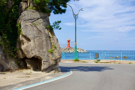 Foto de Condado de Yangyang, Corea del Sur - 30 de julio de 2019: Cerca de Namae Port, una carretera abraza una roca masiva en la base del acantilado, con ramas de pino colgando y el icónico faro rojo en la distancia. - Imagen libre de derechos