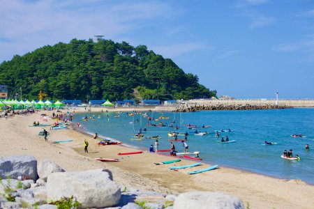 Foto de Condado de Yangyang, Corea del Sur - 30 de julio de 2019: La serena montaña Jukdo, con su observatorio con vistas, establece un pintoresco telón de fondo para los surfistas en el agua y las tablas de surf alineadas en la playa de Ingu. - Imagen libre de derechos