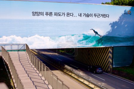Photo for Yangyang County, South Korea - July 30th, 2019: A large billboard spans an overpass, proclaiming "The blue waves of Yangyang County are coming...my heart is pounding," accompanied by an image of a surfer riding the waves, with a switchback walking pa - Royalty Free Image