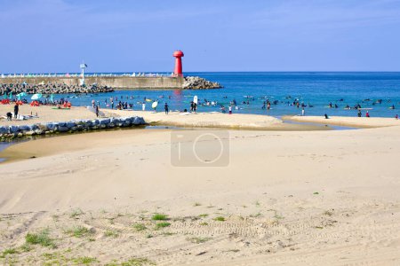 Téléchargez les photos : Comté de Yangyang, Corée du Sud - 30 juillet 2019 : Une journée d'été animée à Gisamun Beach, avec un brise-lames qui s'étend dans la mer de l'Est, coiffé d'un phare rouge, avec des foules et des planches de surf parsemées d'eau. - en image libre de droit