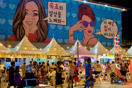 Photo for Sokcho City, South Korea - July 30, 2019: An active night market on Sokcho Beach, under a large building with murals and white popup tents illuminated by overhead lights, as people bustle around the market. - Royalty Free Image