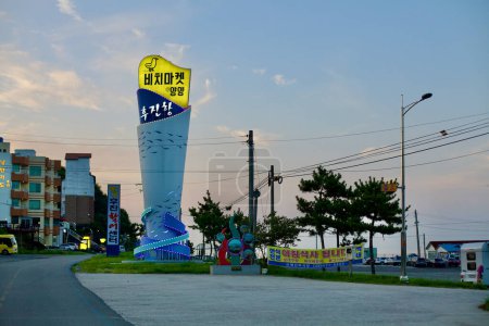 Photo for Yangyang County, South Korea - July 30, 2019: The illuminated entrance sculpture of Hujin Port, a tall cylindrical structure with fish motifs, topped with a duck figure and marked with "Hujin Port" in Korean and "Beach Market @ Yangyang". - Royalty Free Image