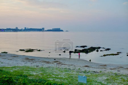 Photo for Yangyang County, South Korea - July 30, 2019: An evocative view of Jeongam Beach after sunset, with two visitors wading near rocks in the shallow water and the distant Oeongchi Port visible in the background. - Royalty Free Image