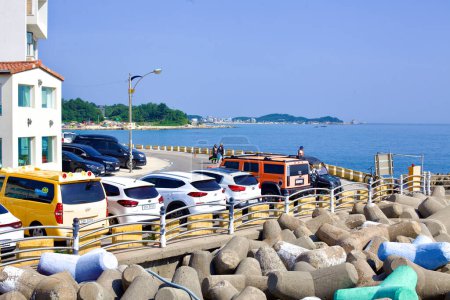 Foto de Condado de Goseong, Corea del Sur - 30 de julio de 2019: En Ayajin Port, el comienzo de un rompeolas con tetrápodos en primer plano, coches estacionados a lo largo de la carretera costera y una cafetería a la izquierda, todo listo en el hermoso telón de fondo de la costa del Mar del Este - Imagen libre de derechos