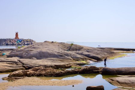 Foto de Condado de Goseong, Corea del Sur - 30 de julio de 2019: Cerca del puerto de Ayajin, un pescador se encuentra en las tranquilas aguas del Mar del Este entre rocas lisas y bajas, con un rompeolas que conduce a un pequeño faro rojo en la distancia. - Imagen libre de derechos