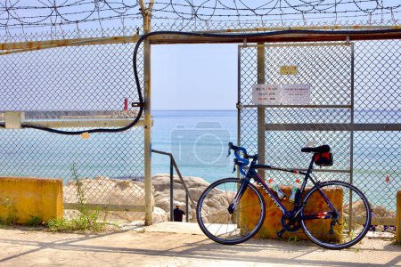 Foto de Condado de Goseong, Corea del Sur - 30 de julio de 2019: Una bicicleta solitaria se apoya contra una puerta abierta en una cerca de alambre de púas en Gonghyeonjin Beach, donde las escaleras invitan a los visitantes a bajar a una playa rocosa debajo. - Imagen libre de derechos