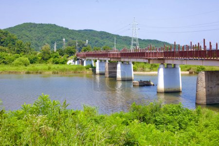Foto de Condado de Goseong, Corea del Sur - 30 de julio de 2019: El histórico puente ferroviario de Bukcheon, que atraviesa un arroyo que desemboca en el Mar del Este, cuenta con una cerca roja a lo largo de su cubierta, con un telón de fondo montañoso. Este puente, destruido durante la Guerra de Corea, permanece como - Imagen libre de derechos