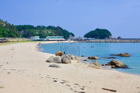 Téléchargez les photos : Comté de Goseong, Corée du Sud - 30 juillet 2019 : La pointe nord sablonneuse de Gonghyeonjin Beach, près du port de Gajin, offre un cadre serein avec une poignée de personnes profitant de l'eau et une colline verte luxuriante visible au loin le long du bord de mer. - en image libre de droit