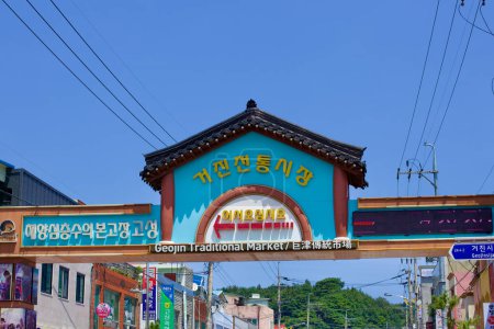 Foto de Condado de Goseong, Corea del Sur - 30 de julio de 2019: Un letrero azul alto marca la entrada al bullicioso mercado tradicional de Geojin a lo largo de la carretera principal a través del puerto de Geojin, invitando a lugareños y visitantes por igual a explorar los puestos vibrantes y productos locales. - Imagen libre de derechos