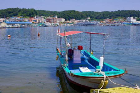 Téléchargez les photos : Comté de Goseong, Corée du Sud - 30 juillet 2019 : Un petit bateau est amarré sur les quais tranquilles de Geojin, flottant sur des eaux calmes et claires. De l'autre côté des eaux, un petit port est visible, sur fond de basses collines verdoyantes, incarnant une livrée côtière paisible. - en image libre de droit