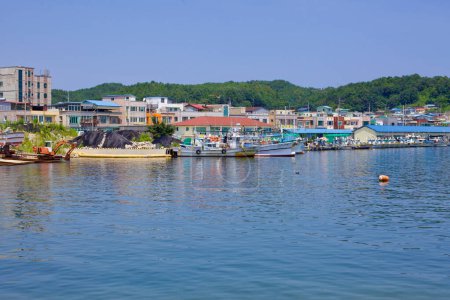 Foto de Condado de Goseong, Corea del Sur - 30 de julio de 2019: El mediodía en el puerto de Geojin durante el verano muestra barcos amarrados a lo largo de los muelles, en un pintoresco telón de fondo de verdes colinas, bajo un cielo azul claro con vibrantes aguas azules que reflejan el ambiente sereno - Imagen libre de derechos