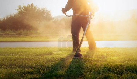 Gros plan d'un travailleur municipal tenant une tondeuse à gazon, coupant l'herbe dans un parc public pendant un lever de soleil vibrant. Entretien de la pelouse à l'aube avec un soleil spectaculaire.
