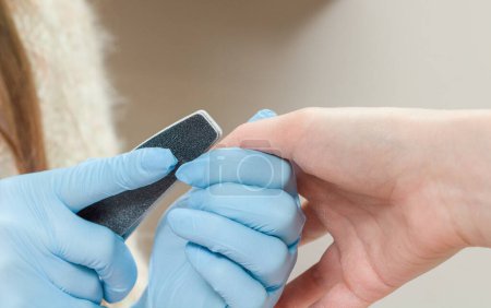 Téléchargez les photos : Manucure en gants bleus lisse un ongle avec une lime à ongles dans un salon de beauté. - en image libre de droit