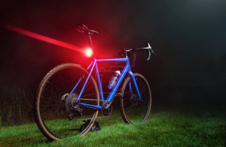 Photo for Blue gravel bike with a bright red flashing light on a green lawn in the dark. Alternative to public transport. - Royalty Free Image