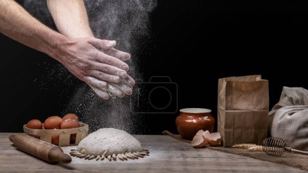 Photo for Baker is cooking bread. Clap hands with flour over the dough. Flour in the air. Cooking homemade food. On the kitchen table are a basket of eggs, a bag of flour, a pot and a rolling pin - Royalty Free Image