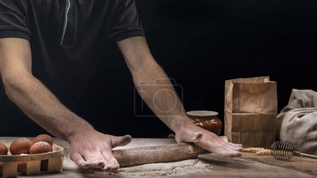 Photo for Man cooks homemade bread in the kitchen. Baker rolls dough with a rolling pin on a wooden table sprinkled with flour - Royalty Free Image