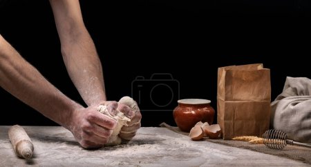 Photo for A baker is cooking bread in the kitchen. A man kneads dough with his hands on a wooden kitchen worktop. Home cooking concept - Royalty Free Image