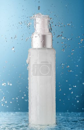 Foto de Botella cosmética con agua que cae y salpica sobre fondo azul. Concepto de frescura y belleza. - Imagen libre de derechos