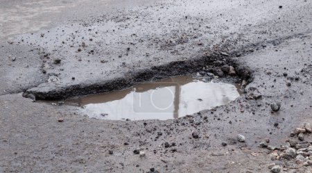 Deep pit on the road with puddle. Damaged asphalt after winter. Road repair.
