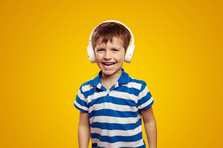 Excited little kid in headphones listening to music and smiling isolated over yellow background, wearing blue striped polo shirt.