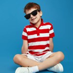 Vertical photo of adorable boy in casual outfit and trendy sunglasses feels happy sitting against the blue background and looking at the camera.