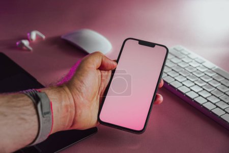 Hand holding smartphone with blank screen. Electronic devices on pink pastel background.