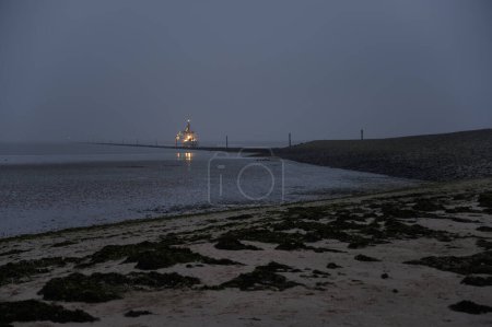 Ferry arriving from the island of Spiekeroog at dusk in Neuharlingersiel, Germany with two more water taxi's lights visible in the distance and piles of sea shells and algae visible due to low tide