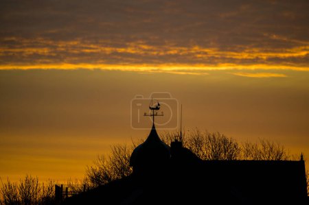 Weathervane on top of a building with a fishing boat as wind direction finder and cardinal directions indicated below during a spectacular orange sunrise.