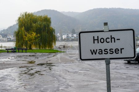 A sign in German saying "Hochwasser" meaning high water or flooding with the Rhine river and a partially flooded parking lot in out of focus background.