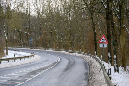 Serpentinenstraße in einem verschneiten Wald mit einem rutschigen Straßenschild und schmutzigem, matschigen Schnee auf der Seite unterhalb der Absperrung.
