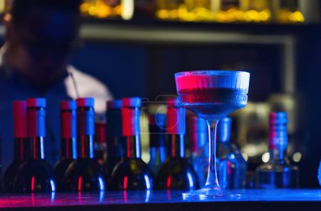 Téléchargez les photos : Cocktail sur comptoir de bar avec bouteilles et fond bokeh foncé, éclairé par un néon rouge et bleu. - en image libre de droit