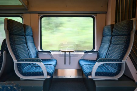 Téléchargez les photos : Sièges bleus vides dans le train de voyageurs avec un feuillage flou à l'extérieur de la fenêtre pendant la journée. - en image libre de droit