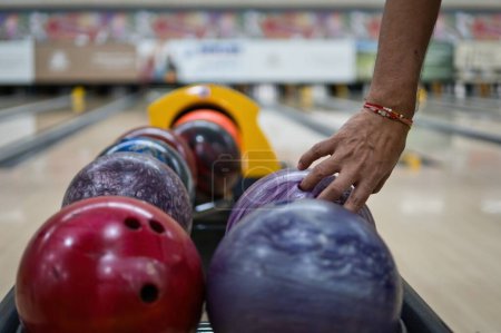 Hand picking up bowling balls in return machine at bowling alley. Lanes and pins in blurred background.