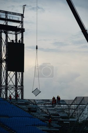 Téléchargez les photos : Travailleurs munis de casquettes et de gilets de sécurité installant des sièges sur la tribune, couverts et nuageux. - en image libre de droit