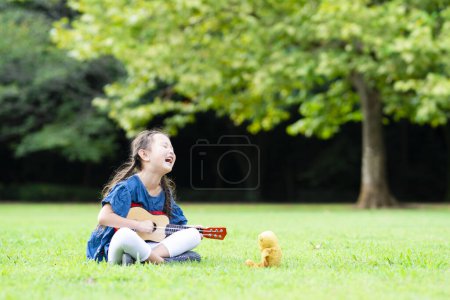 Téléchargez les photos : Petite fille asiatique avec ours en peluche jouant ukulele dans le parc. - en image libre de droit
