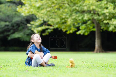 Téléchargez les photos : Petite fille asiatique avec ours en peluche jouant ukulele dans le parc. - en image libre de droit